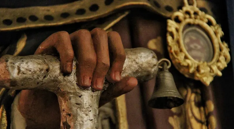 La mano della statua stretta sul bastone a forma di Tau, con la campanella appesa
